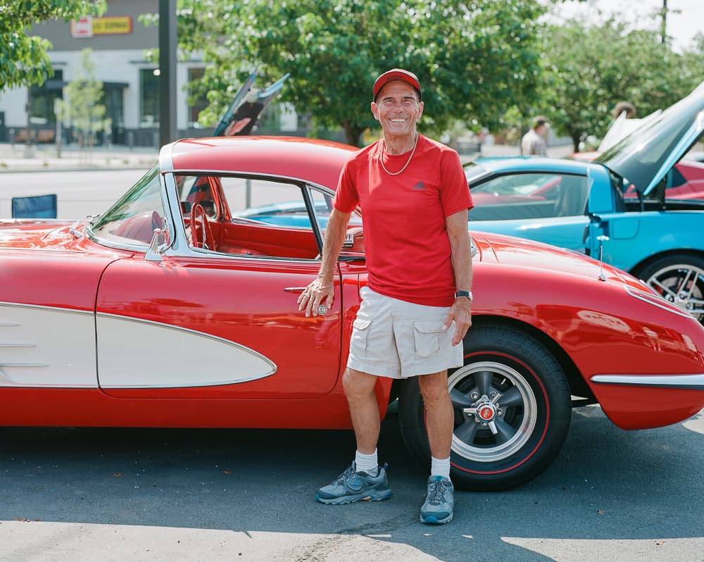 Man in a red shirt and cap leaning against a red and white classic Corvette at a car show