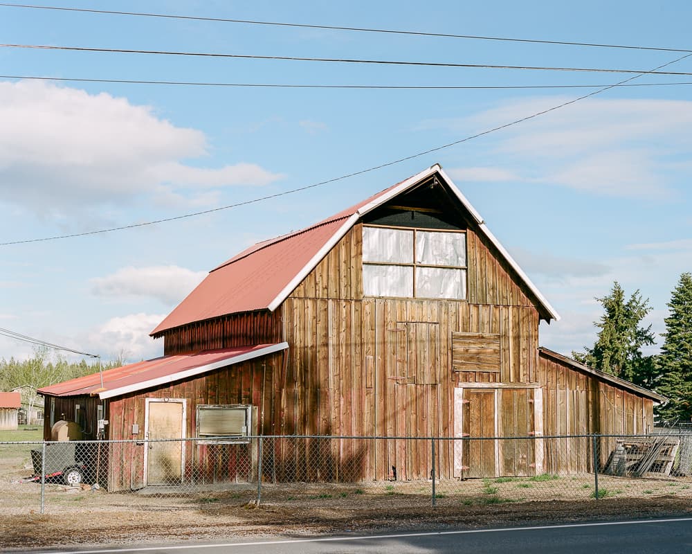 Weathered wooden barn with a red metal roof behind a chain-link fence in evening light