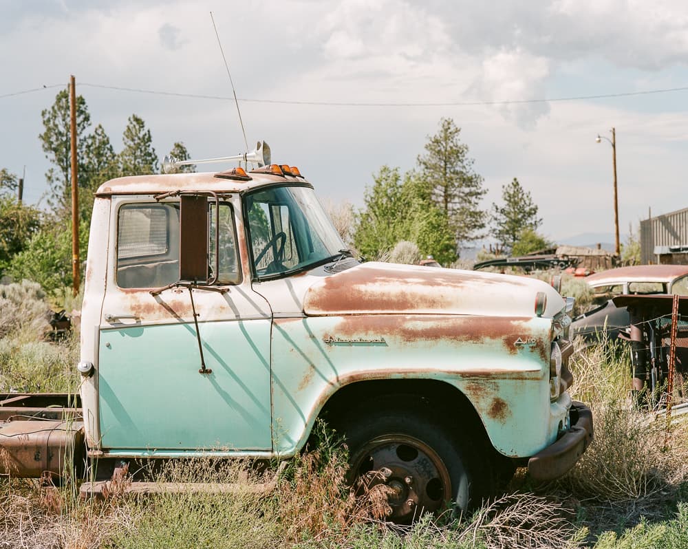 Rusted turquoise farm truck sitting in overgrown grass under a cloudy high-desert sky