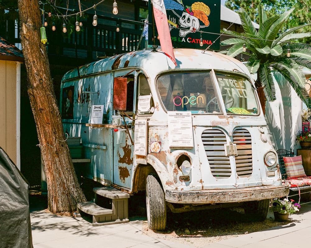 Weathered vintage step van converted to a food truck with a neon open sign, parked beneath a palm tree