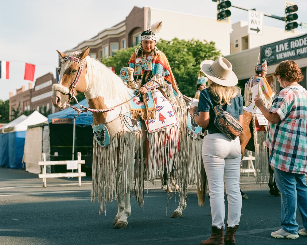 Rider in traditional regalia on a horse decorated with fringe, greeting onlookers at a downtown rodeo parade