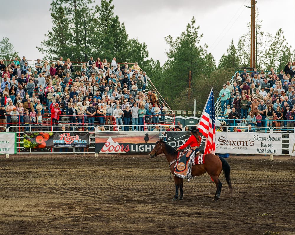 Rider carrying an American flag on horseback in front of a packed rodeo grandstand